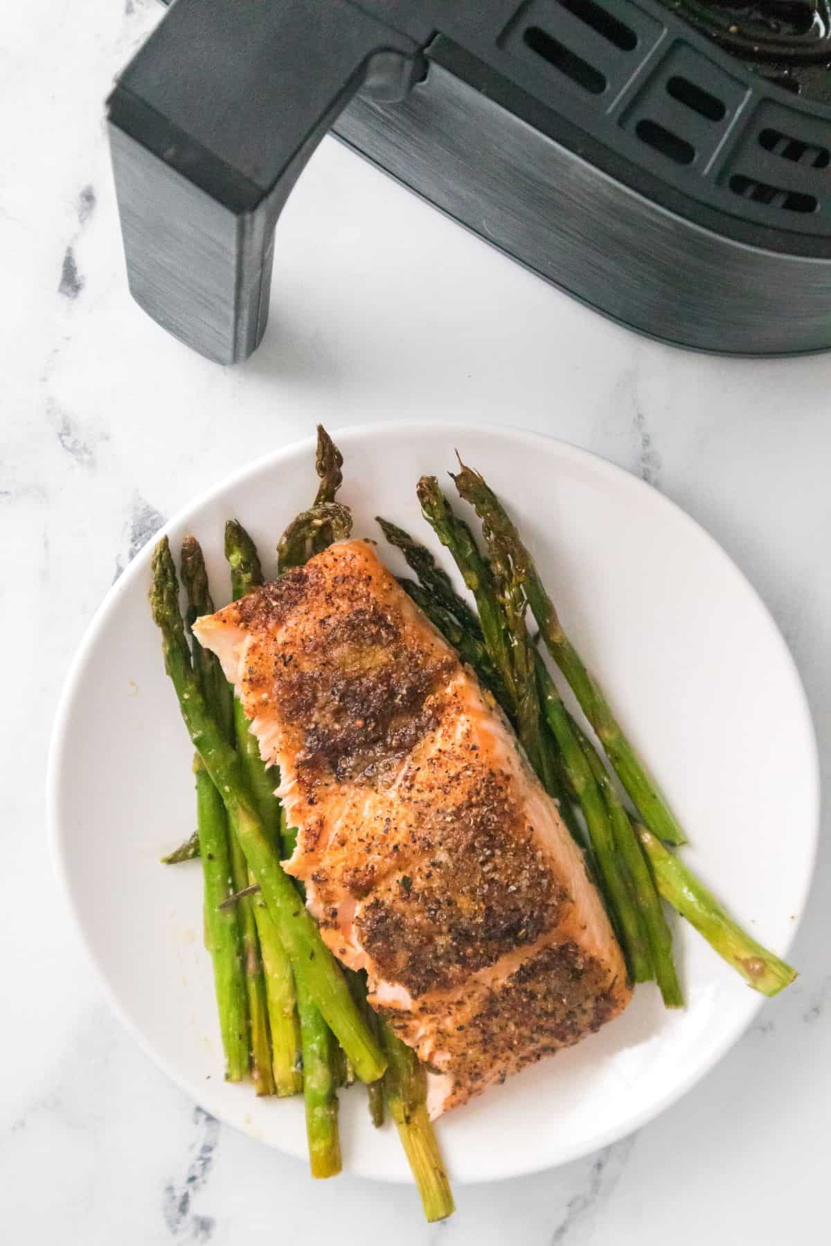 A plate with a cooked fish and featuring tender roasted vegetables sits next to an air fryer on a white surface.