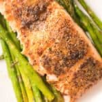 A seasoned, baked salmon fillet is elegantly served on a bed of roasted asparagus spears, showcasing the perfect harmony of salmon and asparagus on a pristine white plate.