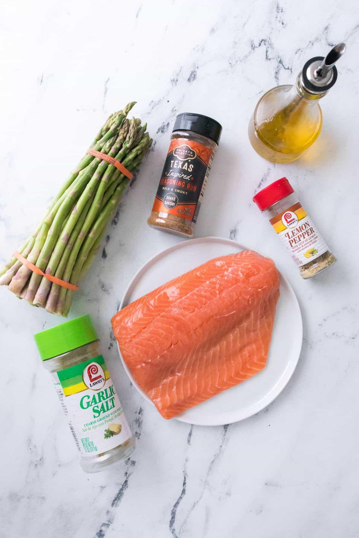 Raw salmon fillets and a bundle of fresh asparagus rest on a plate, accompanied by garlic salt, lemon pepper, Texas steak rub, and a bottle of olive oil on a marble surface—perfect for creating a flavorful salmon and asparagus meal.
