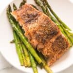 A delicious serving of Salmon and Asparagus features a cooked salmon fillet seasoned with pepper, sitting atop perfectly roasted asparagus spears on a white plate.