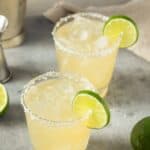 Two glasses of Classic Margarita with salted rims, ice, and lime slices sit on a gray surface. A fresh lime and metal jigger are nearby.