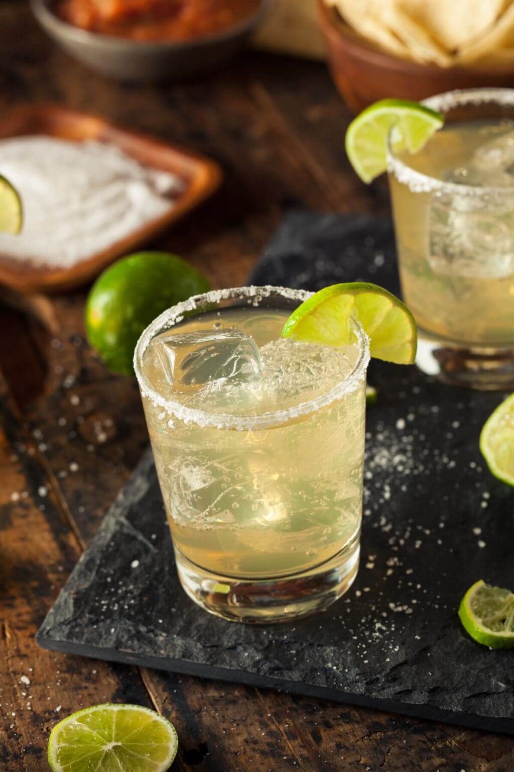 A glass with a salted rim, ice cubes, and a lime wedge garnish sits on a dark surface, surrounded by scattered lime slices and salt.