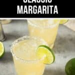 A glass of Classic Margarita with a salted rim and a lime slice garnish sits on a gray surface, with another margarita and lime halves in the background.
