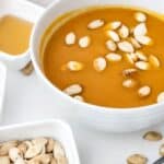 A bowl of orange soup, reminiscent of Panera Autumn Squash Soup, topped with pumpkin seeds and surrounded by a small bowl of pumpkin seeds, a spoon, and a small dish of light-colored sauce.