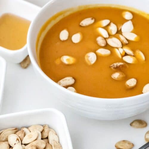 A bowl of orange soup, reminiscent of Panera Autumn Squash Soup, topped with pumpkin seeds and surrounded by a small bowl of pumpkin seeds, a spoon, and a small dish of light-colored sauce.