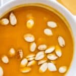A white bowl filled with soup, reminiscent of Panera Autumn Squash Soup, is topped with a scattering of pumpkin seeds and shown on a white surface.
