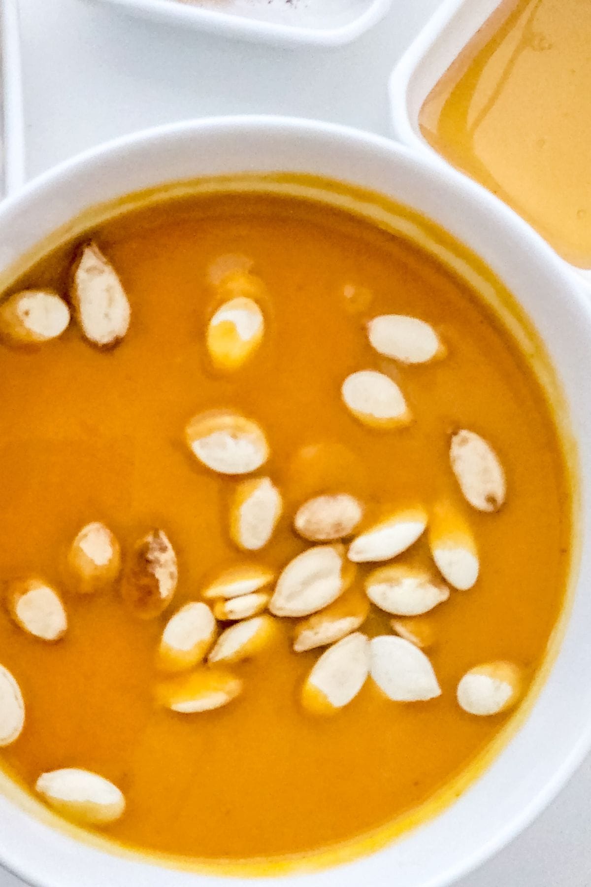 A white bowl filled with soup, reminiscent of Panera Autumn Squash Soup, is topped with a scattering of pumpkin seeds and shown on a white surface.