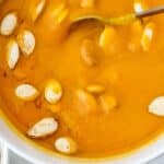A bowl of orange Panera Autumn Squash Soup garnished with pumpkin seeds, with a spoon resting in the soup.