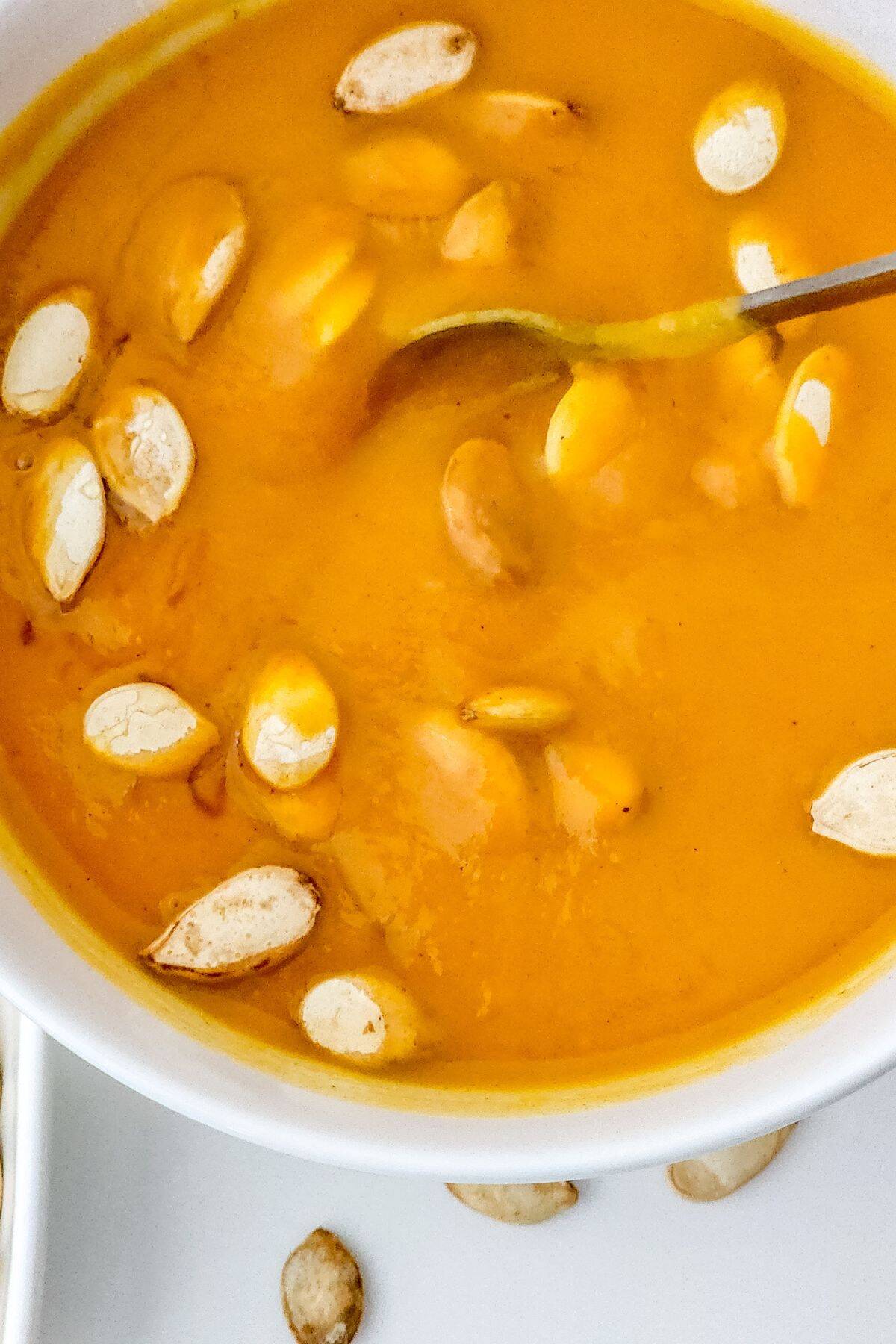 A bowl of orange Panera Autumn Squash Soup garnished with pumpkin seeds, with a spoon resting in the soup.