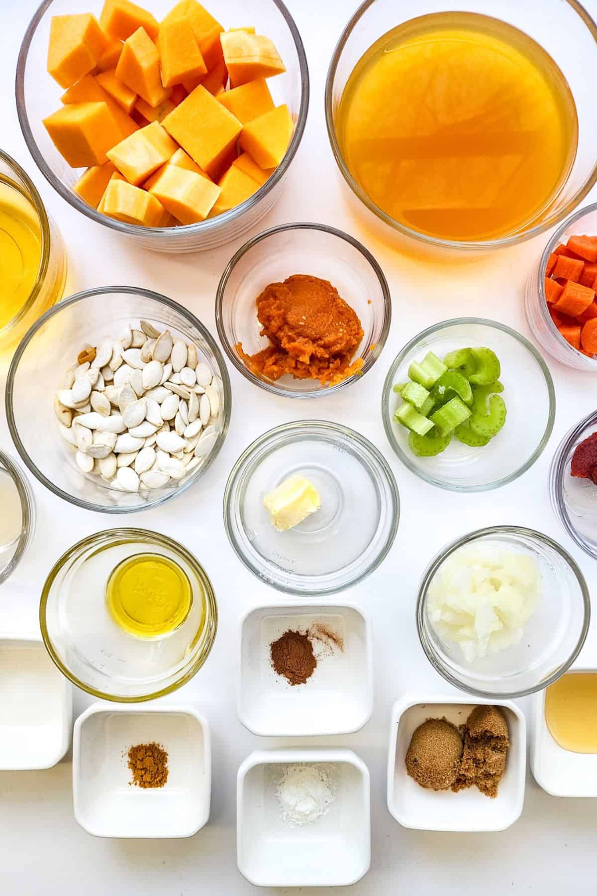 Assorted glass bowls and ramekins containing cubed squash, vegetable broth, pumpkin puree, chopped carrots, celery, pumpkin seeds, butter, onion, oil, and spices—everything needed.