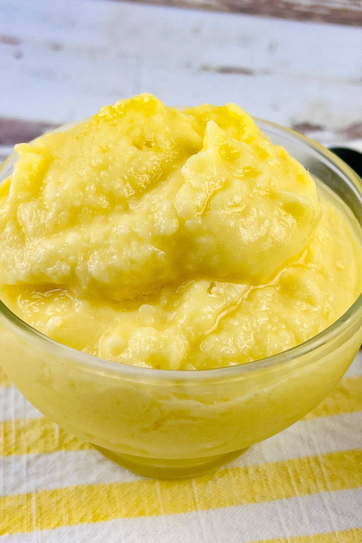 A clear glass bowl filled with thick, smooth, yellow Dole Pineapple Whip sits on a striped yellow and white cloth.