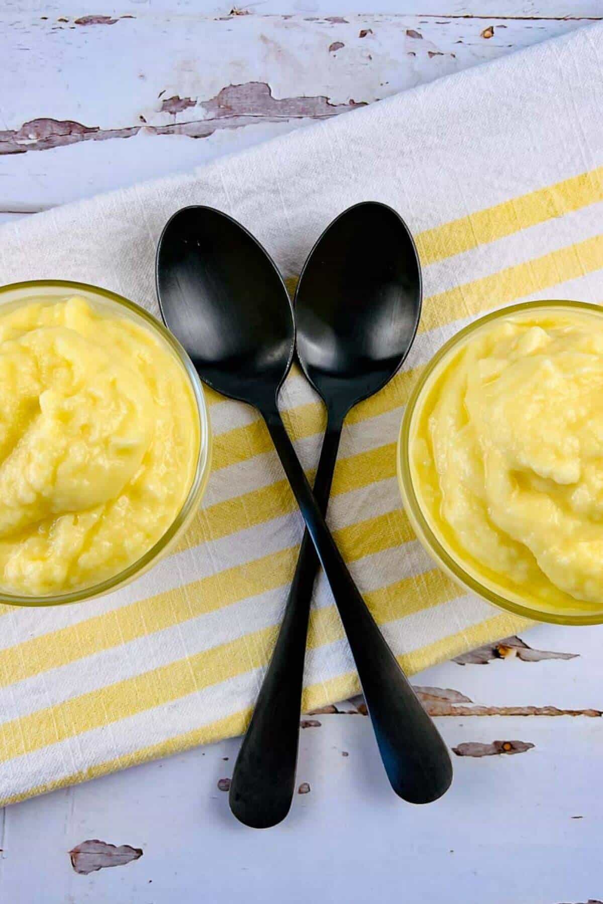 Two bowls of creamy yellow pudding, reminiscent of Dole Pineapple Whip, sit on a yellow and white striped cloth with two black spoons crossed in the center.