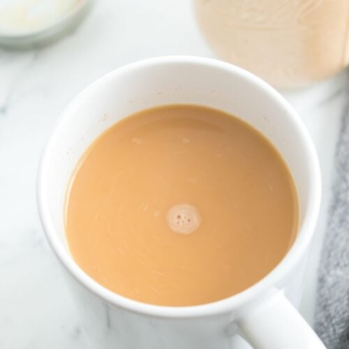 A white mug filled with light brown coffee sits on a marble surface next to a jar with a lid and a gray cloth, featuring rich notes of Homemade Pumpkin Spice Creamer.