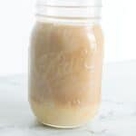 A glass mason jar filled with a creamy beige smoothie—made extra cozy with Homemade Pumpkin Spice Creamer—sits on a white marble surface against a plain white background.