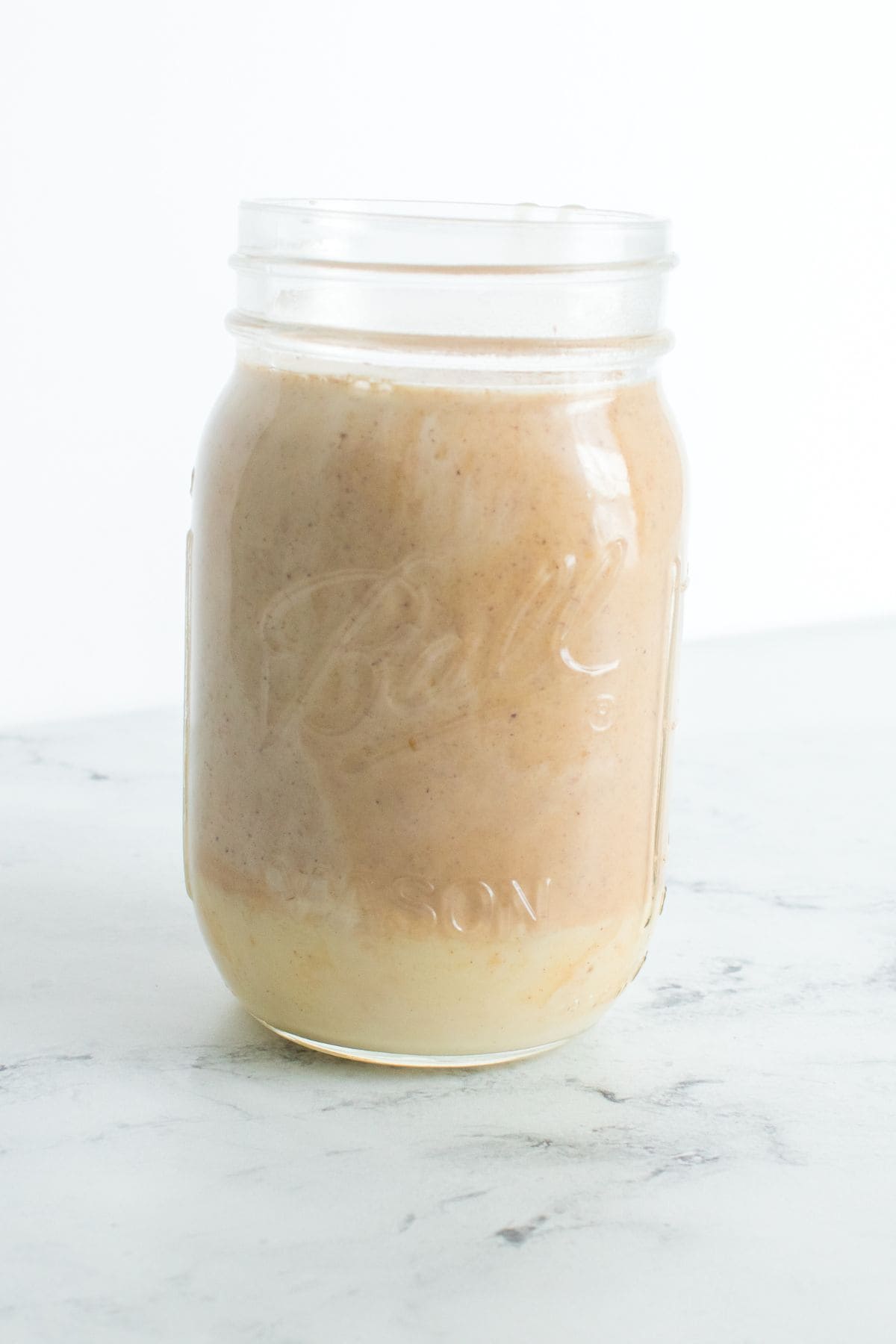 A glass mason jar filled with a creamy beige smoothie—made extra cozy with Homemade Pumpkin Spice Creamer—sits on a white marble surface against a plain white background.