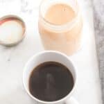 A mug of black coffee sits next to an open glass jar on a white marble surface.