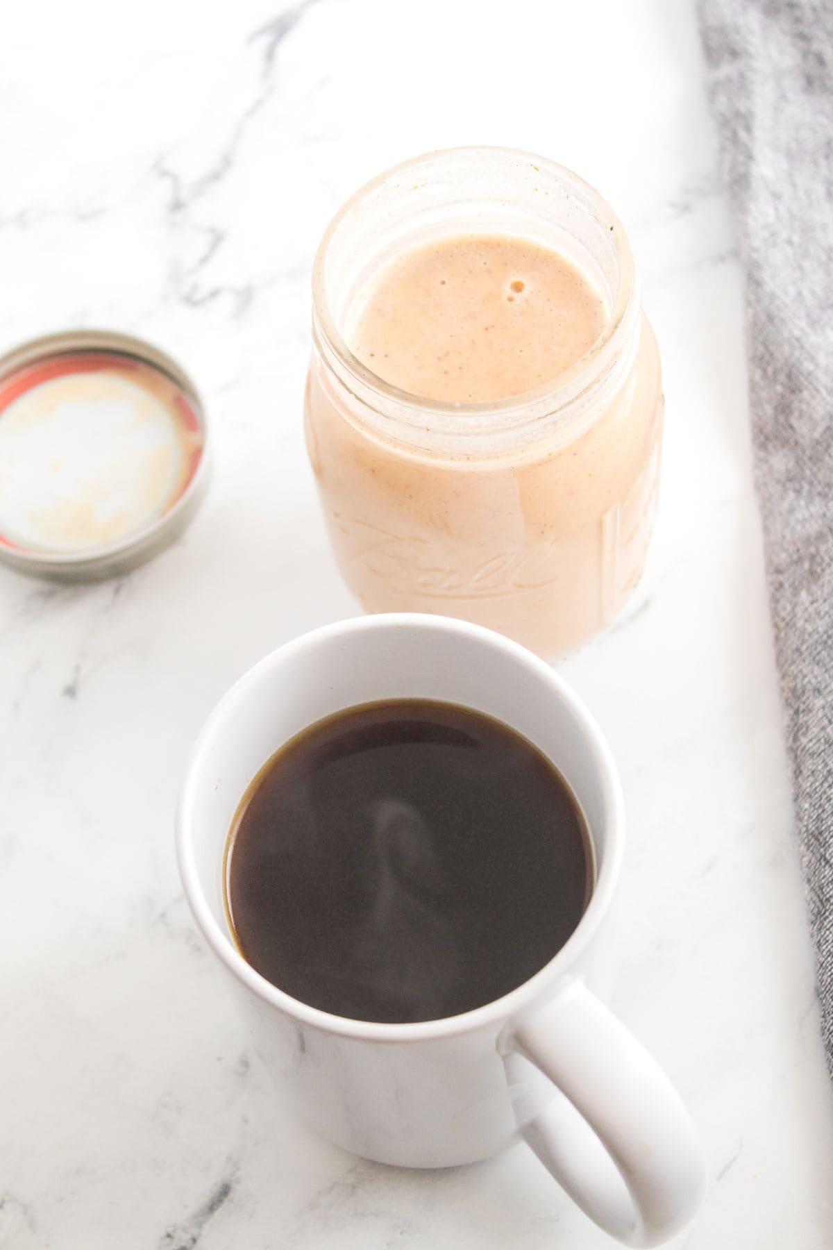 A mug of black coffee sits next to an open glass jar on a white marble surface.