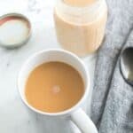 A white mug filled with light brown coffee sits on a marble surface near an open jar of Homemade Pumpkin Spice Creamer, a spoon, and a gray napkin.