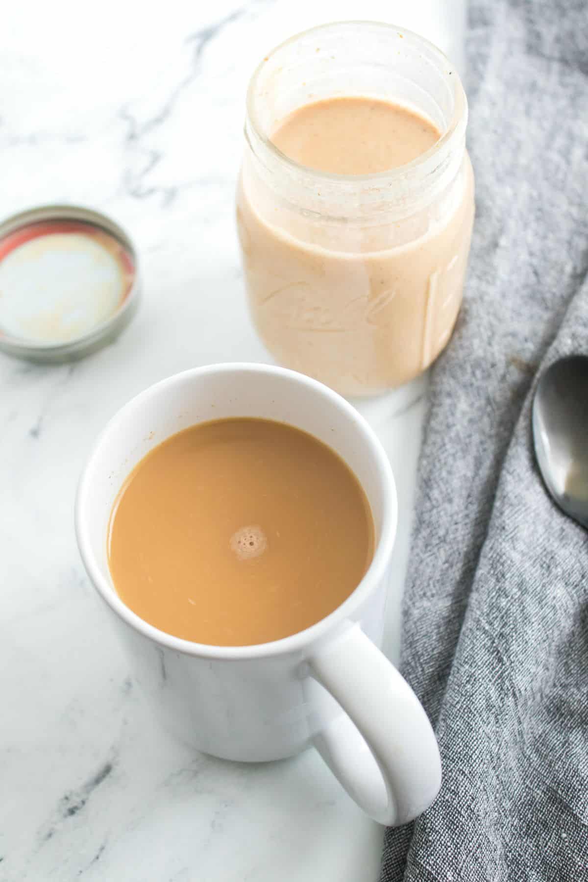 A white mug filled with light brown coffee sits on a marble surface near an open jar a spoon, and a gray napkin.