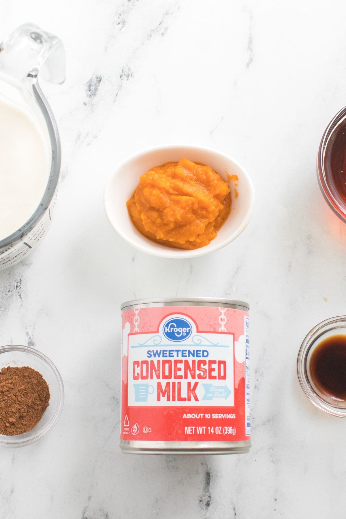 Ingredients: a can of sweetened condensed milk, a bowl of pumpkin puree, a measuring cup of milk, ground spices, vanilla extract, and syrup arranged on a white surface.
