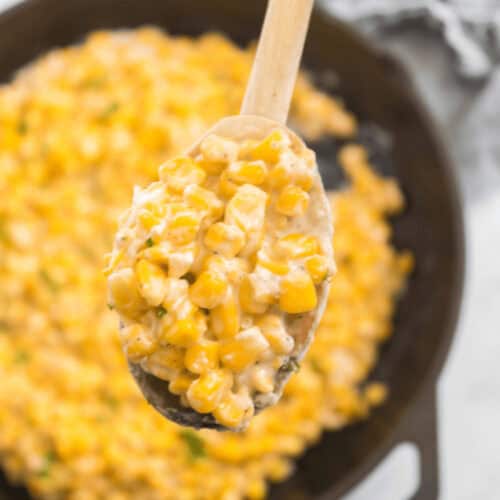 A wooden spoon holds a serving of creamy Honey Butter Skillet Corn mixture above a cast iron skillet filled with the same dish.