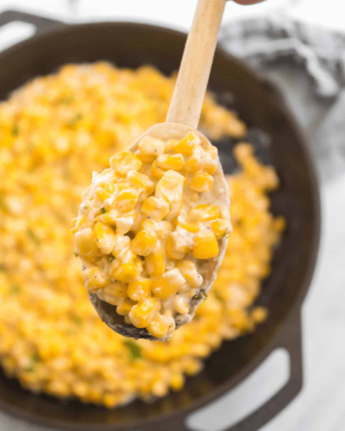 A wooden spoon holds a serving of creamy Honey Butter Skillet Corn mixture above a cast iron skillet filled with the same dish.