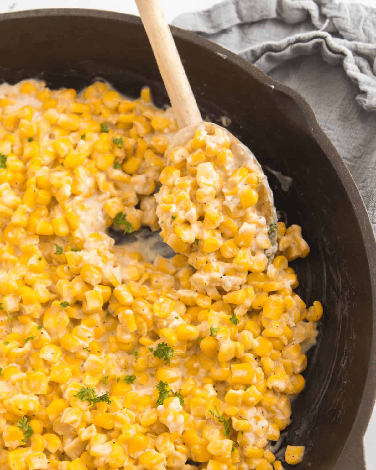 Cooked Honey Butter Skillet Corn in a skillet with a wooden spoon, garnished with chopped parsley, next to a folded gray cloth napkin.