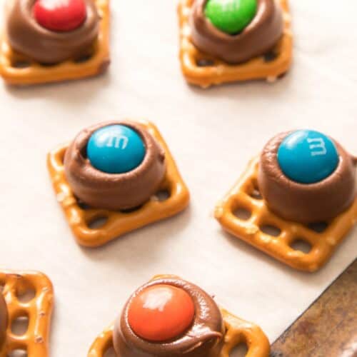 Square pretzels with Hershey Kisses and colorful M&M's are arranged on a parchment-lined baking sheet, creating a sweet and salty treat.