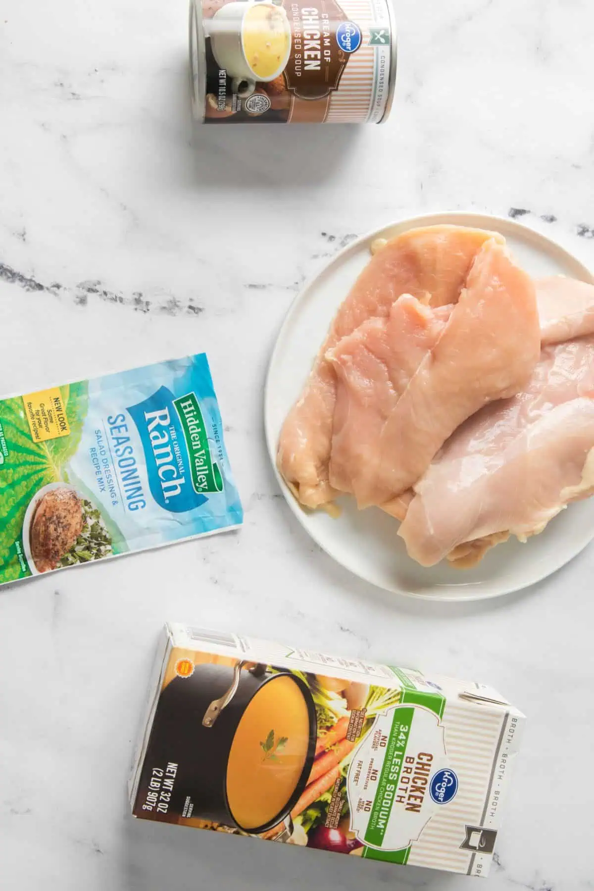 Raw chicken breasts on a white plate, a can of cream of chicken soup, ranch seasoning, and chicken broth on a white marble surface.