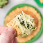 A person holding a cracker topped with spinach and cheese.
