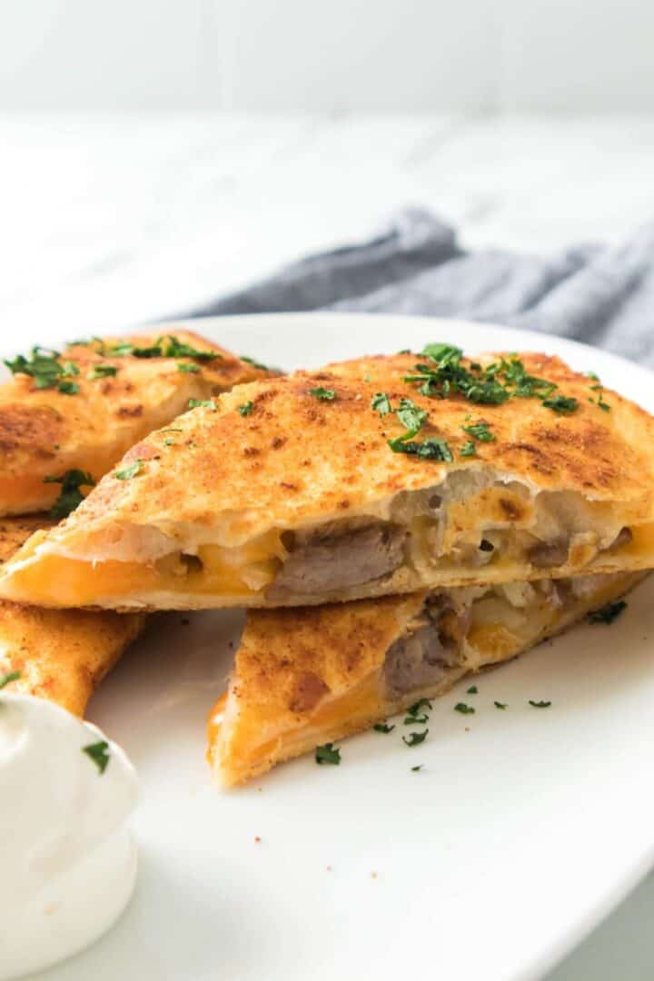A close-up of sliced Steak Quesadillas filled with sausage and melted cheese, garnished with chopped herbs, and served with a dollop of sour cream on a white plate.