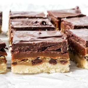 Twix Cookie Bars with a shortbread base, visible chocolate chips, a gooey caramel layer, and thick chocolate topping, cut into squares and arranged on white parchment paper.