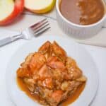 A slice of dessert with a drizzle on top sits on a white plate, accompanied by a fork, a halved apple, and a ramekin in the background.