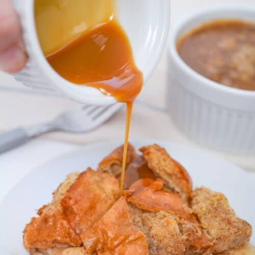A hand pours rich caramel sauce from a small white pitcher onto a warm Bread Pudding with Bourbon Sauce, served on a white plate.