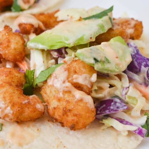 Close-up of a taco filled with Boom Boom Shrimp, avocado slices, coleslaw mix, and drizzled sauce on a corn tortilla.