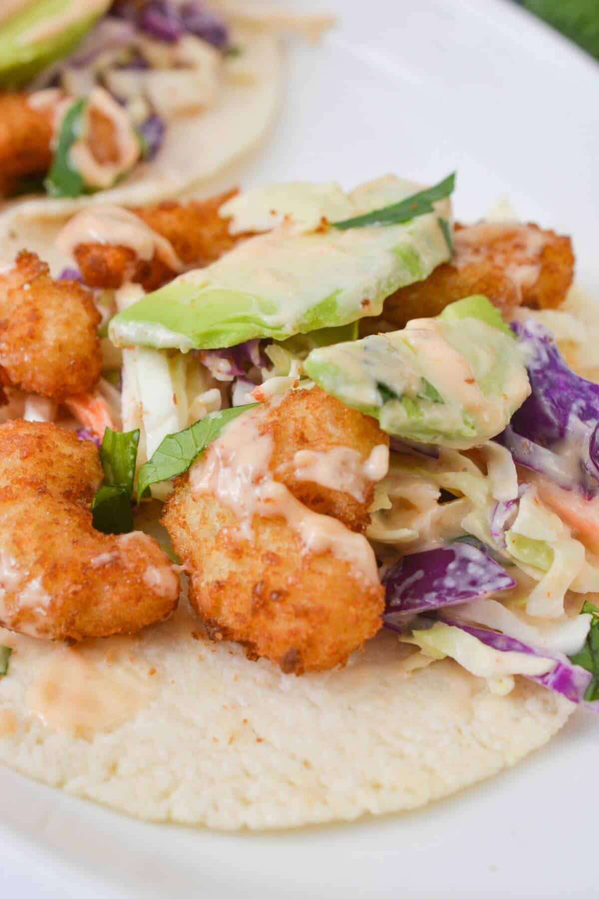 Close-up of a taco filled with Boom Boom Shrimp, avocado slices, coleslaw mix, and drizzled sauce on a corn tortilla.