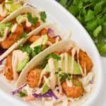 Fish tacos topped with Boom Boom Shrimp, avocado slices, cabbage slaw, and cilantro, all drizzled with a creamy sauce on a white platter. Fresh cilantro and a bowl of slaw in the background add to the vibrant presentation.