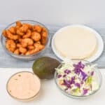 Bowls of Boom Boom Shrimp, tortillas, cabbage slaw, and sauce are elegantly displayed on a table with a whole avocado nearby.