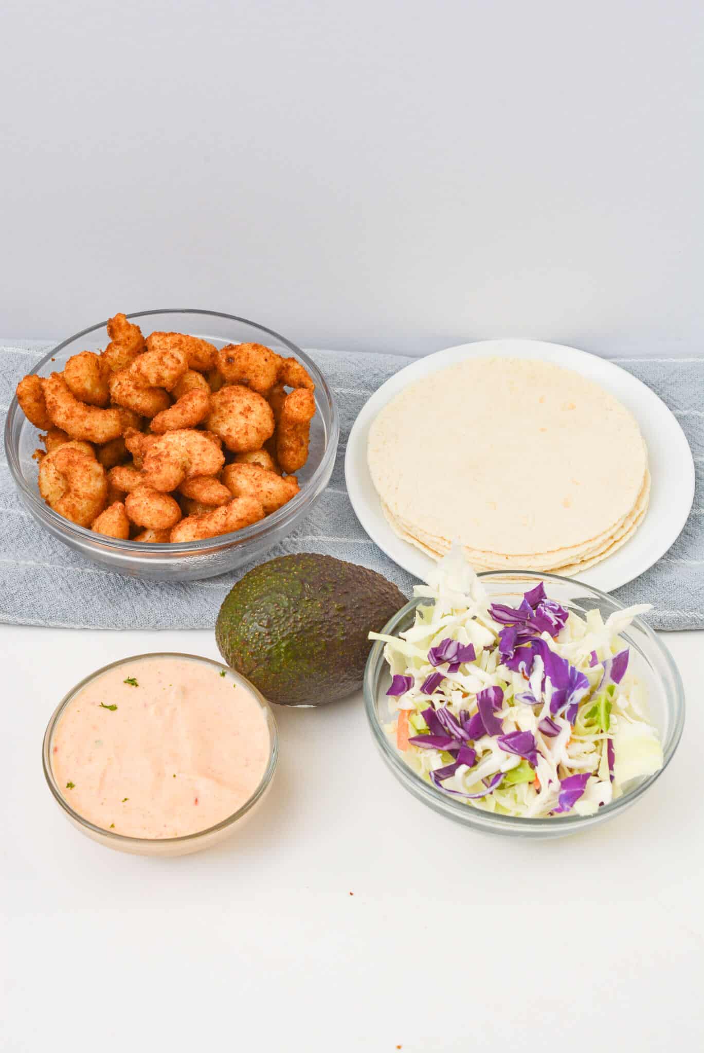 Bowls of Boom Boom Shrimp, tortillas, cabbage slaw, and sauce are elegantly displayed on a table with a whole avocado nearby.