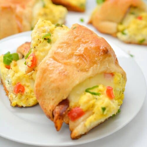 A breakfast Crescent Roll Ring stuffed with scrambled eggs, diced vegetables, bacon, and melted cheese sits on a white plate, with more golden croissants in the background.