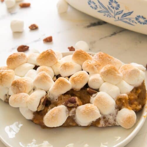 A slice of Candied Sweet Potatoes with Canned Sweet Potatoes casserole, topped with toasted marshmallows and pecans, sits on a white plate, with a baking dish and scattered mini marshmallows in the background.