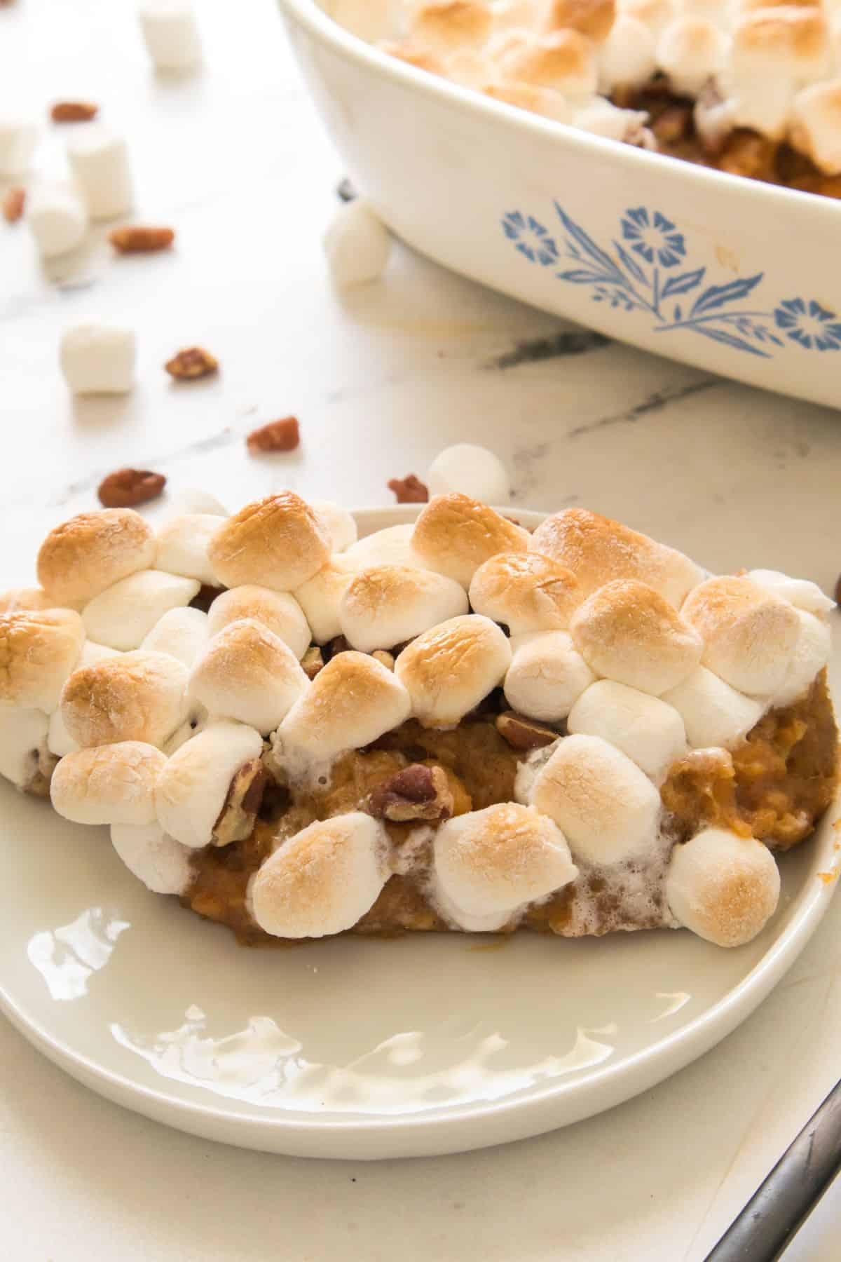 A slice of Candied Sweet Potatoes with Canned Sweet Potatoes casserole, topped with toasted marshmallows and pecans, sits on a white plate, with a baking dish and scattered mini marshmallows in the background.