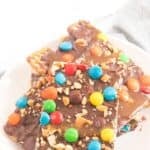 A white plate with Caramel Pretzel Bark, topped with colorful candy-coated chocolates, pretzels, and chopped nuts.