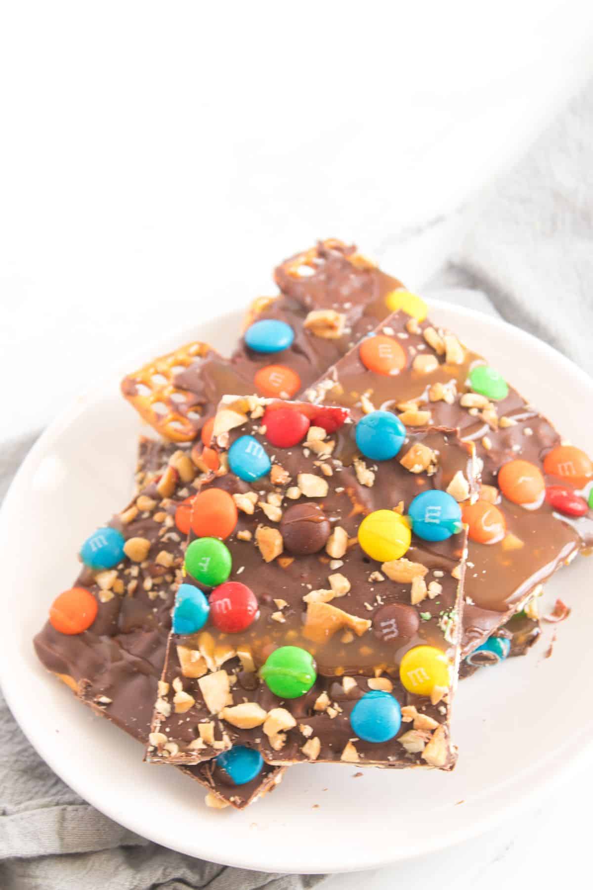 A plate of chocolate-covered toffee bars, similar to Caramel Pretzel Bark, topped with colorful candy-coated chocolates and chopped nuts.