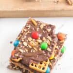 A stack of pieces topped with pretzels, M&M candies, and chopped nuts on a white surface.