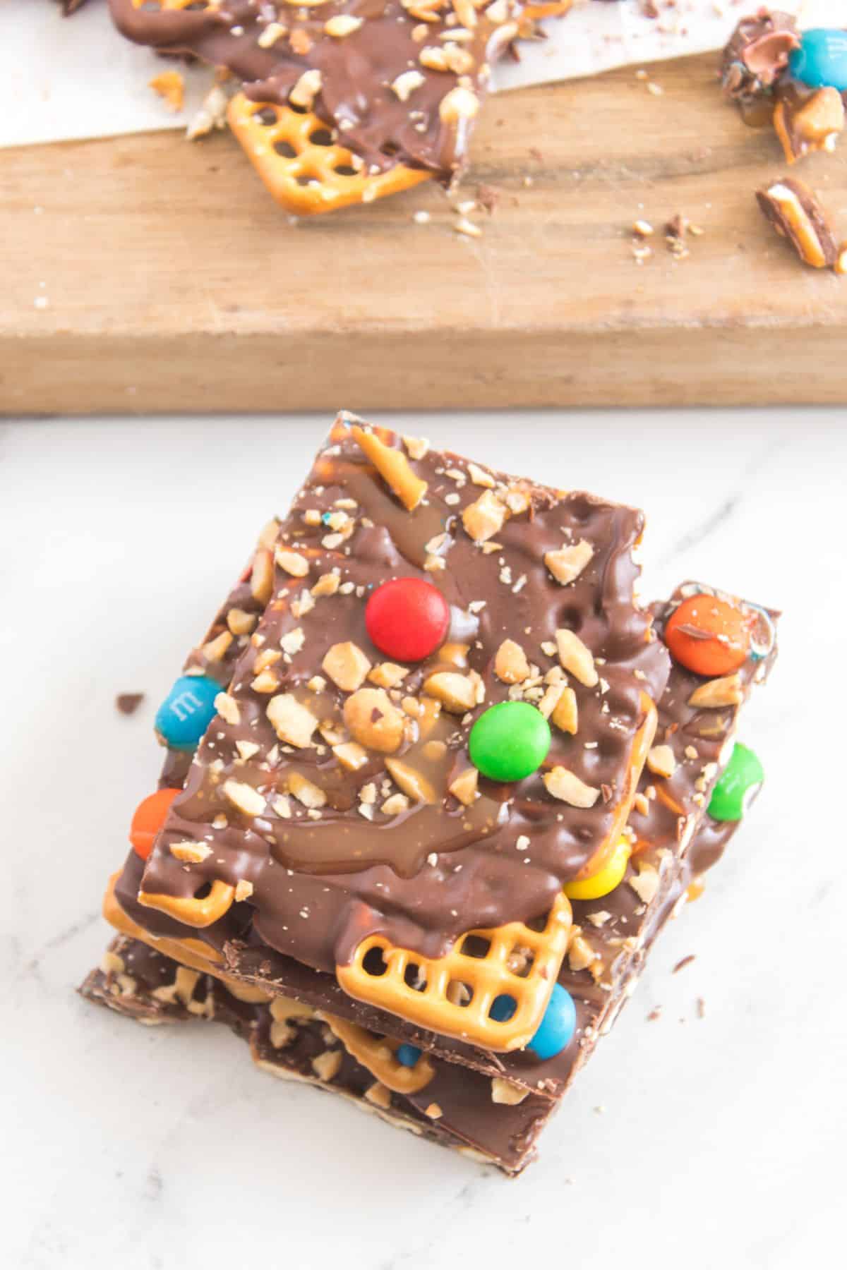 A stack of pieces topped with pretzels, M&M candies, and chopped nuts on a white surface.