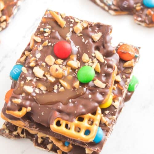 Caramel Pretzel Bark topped with pretzels, peanuts, and colorful candy-coated chocolates on a white surface.