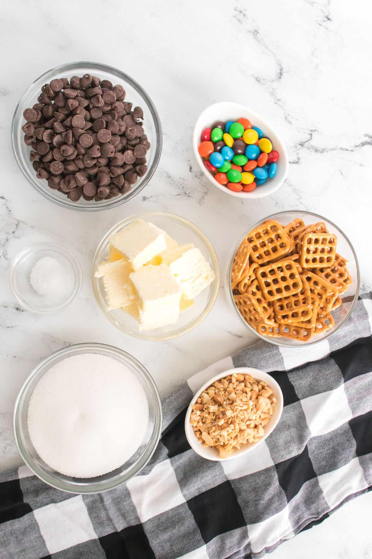Bowls containing chocolate chips, colorful candies, butter, chopped nuts, sugar, salt, and pretzels are arranged on a marble surface next to a black and white checkered cloth—ready to make delicious Caramel Pretzel Bark.