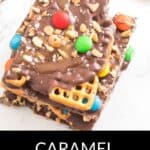 Three pieces of Caramel Pretzel Bark, topped with mini pretzels, peanuts, colorful candy-coated chocolates, and a layer of chocolate, are stacked on a white surface.
