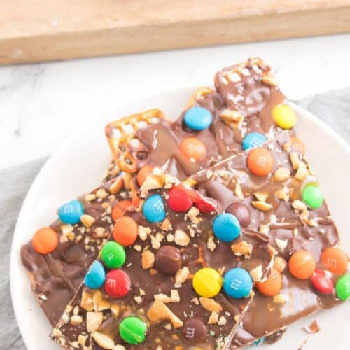 Caramel Pretzel Bark pieces topped with colorful candy-coated chocolates and chopped nuts are arranged on a white plate, with more pieces on a wooden board in the background.